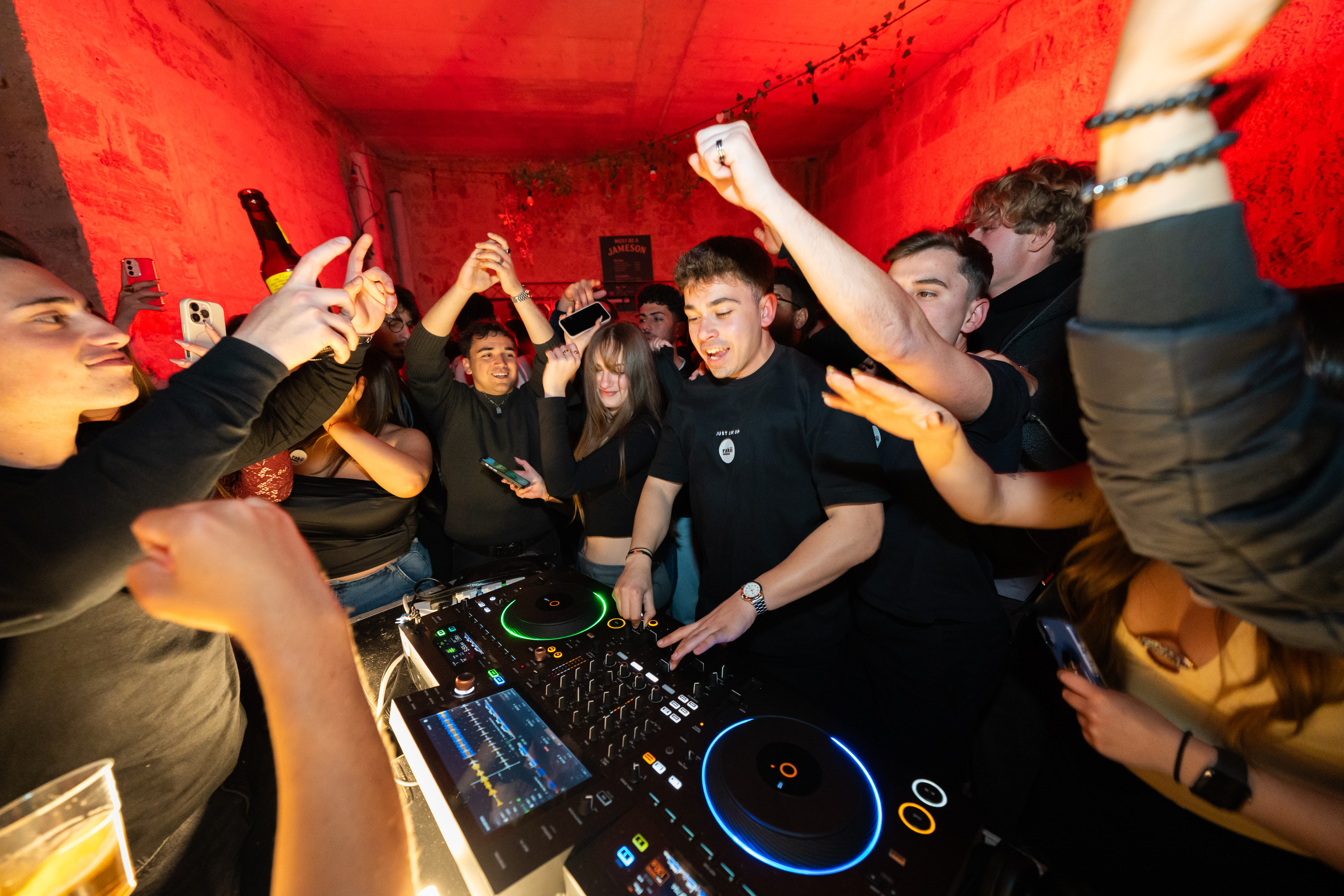 DJ surrounded by crowd under red lighting at Rakii